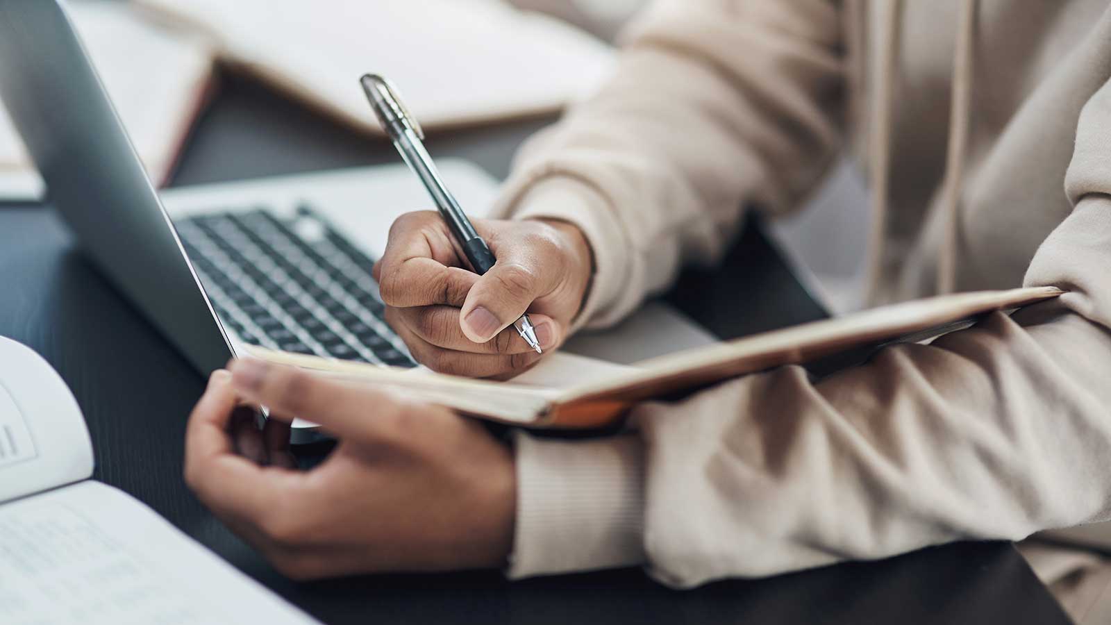 Student taking notes in notebook next to a laptop computer