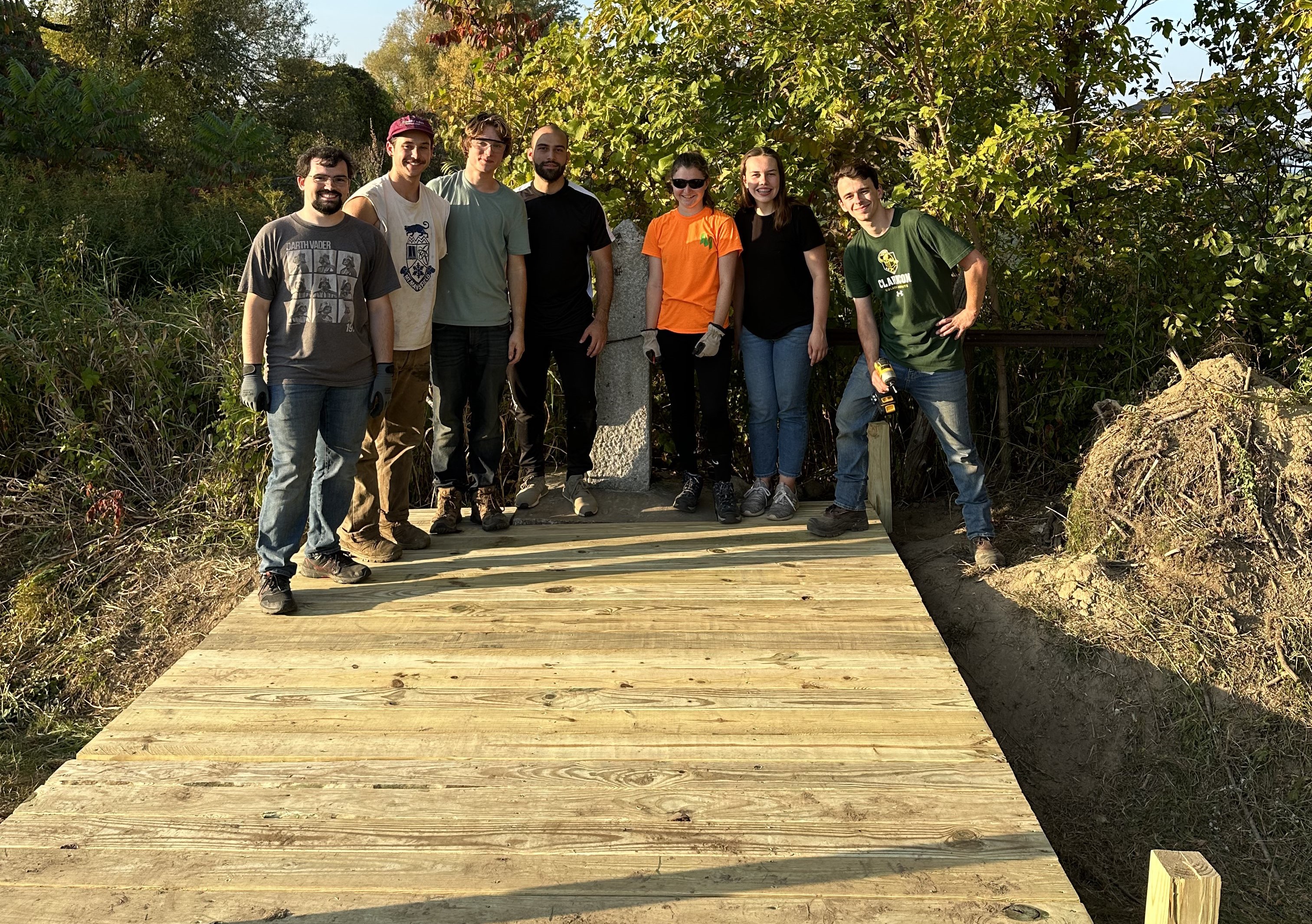 Alt Text: Members of the Timber Bridge SPEED team stand on the bridge they built that leads to a border marker along the US Canada border.