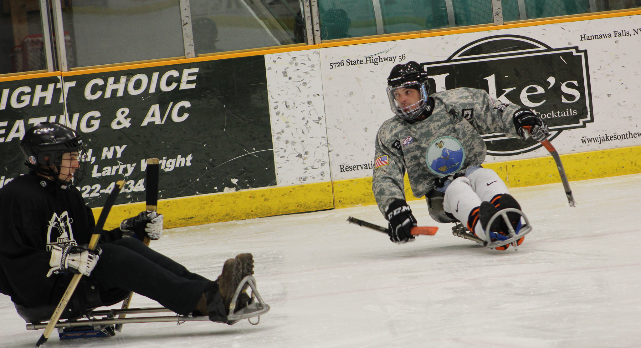 A sled hockey player in a camouflage jersey pushes forward against an opponent in a black jersey