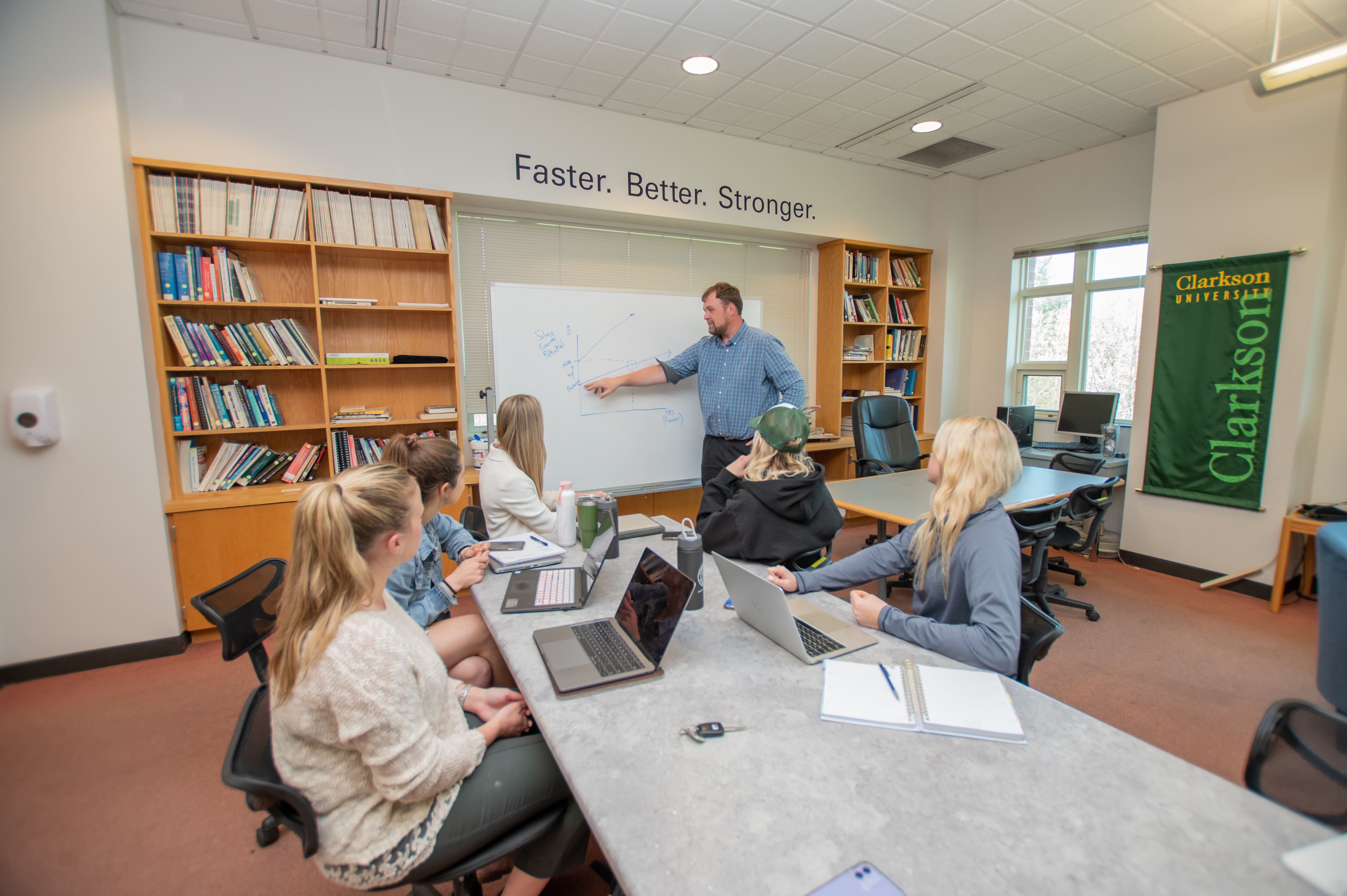 three students sit at a table with their laptops open as a professor points to a white board depicting a line graph with upward projections.