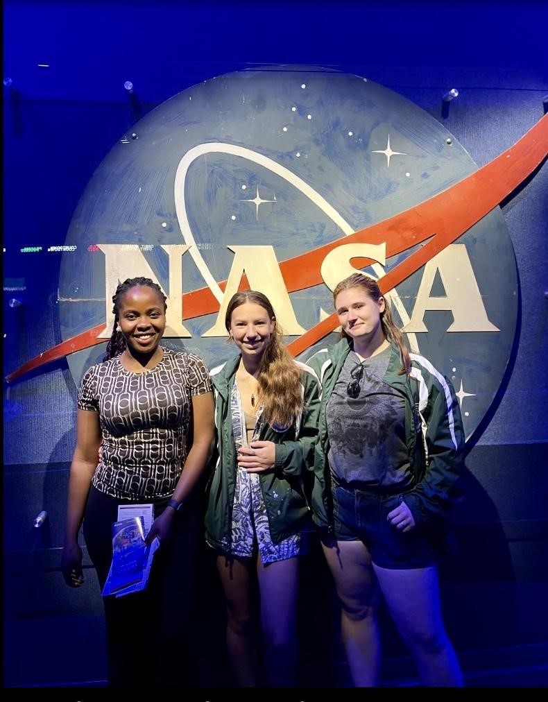 Two students and their teacher pose in front of a large NASA logo. 