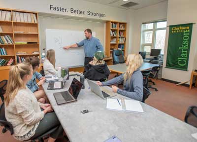 Five students sitting at a long table with notebook computers, while a teacher stands by them at a wall-mounted whiteboard.