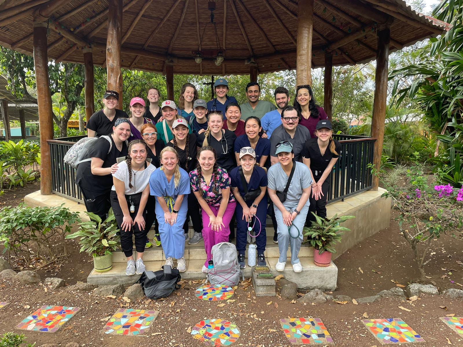 A group of students and medical professionals stand in three rows, touching their knees as they pose for a photo wearing scrubs on the steps of a wooden gazebo.