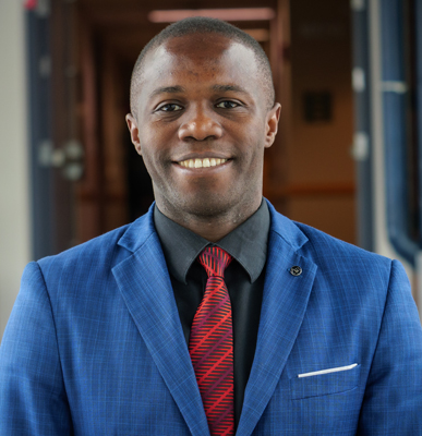 Chest-up portrait of Bebonchu Atems in blue sports coat, black shirt and red tie with blurred corridor in background