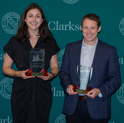 Victoria Ballestero (in black dress) and Bryan Greene (in blue sports jacket, black pants and a blue open-collared shirt) pose against a Clarkson backdrop holding their Woodstock Awards.