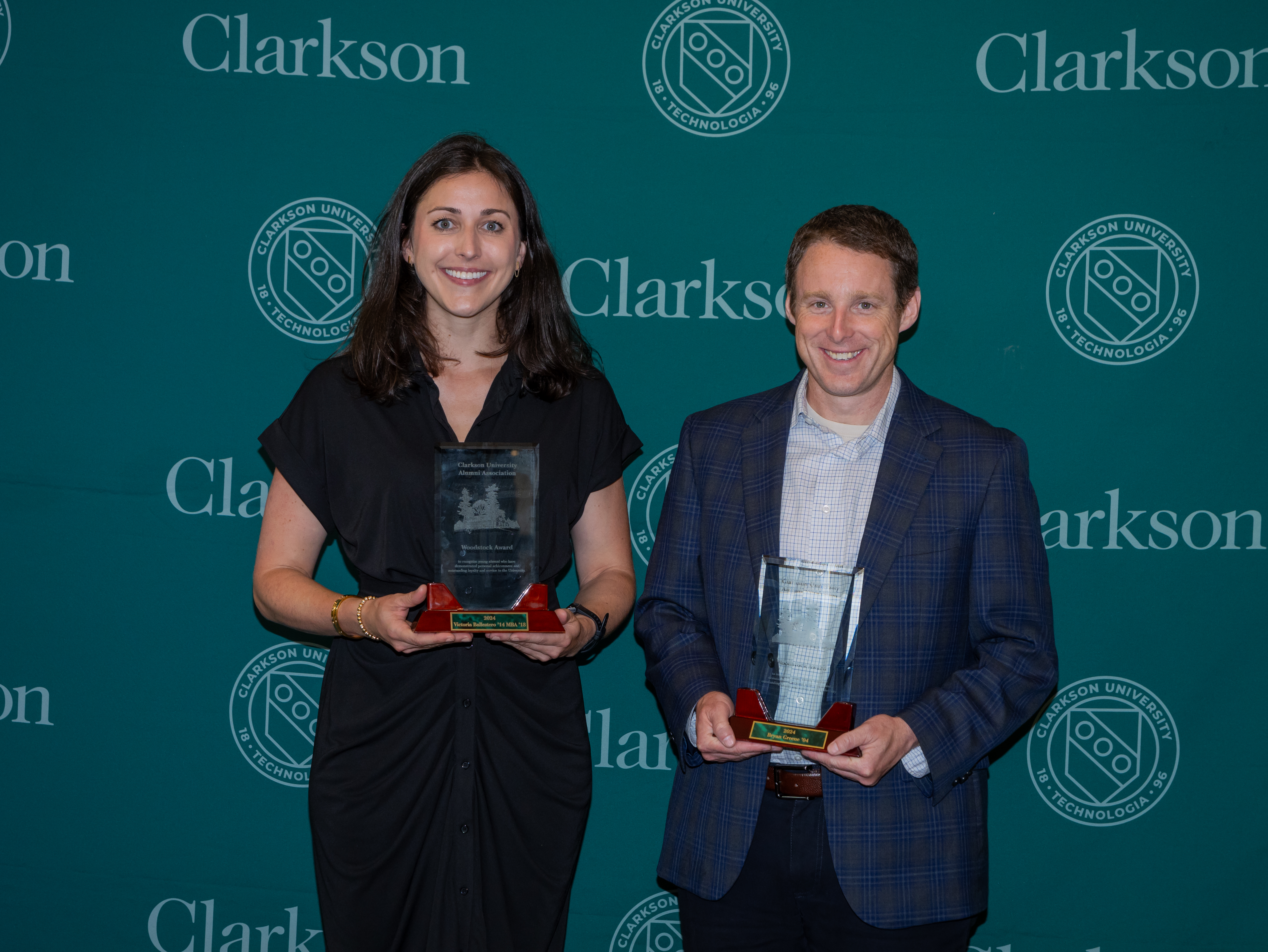 A woman and man stand next to each other posing with crystal plaques in front of a green Clarkson University branded media backdrop.