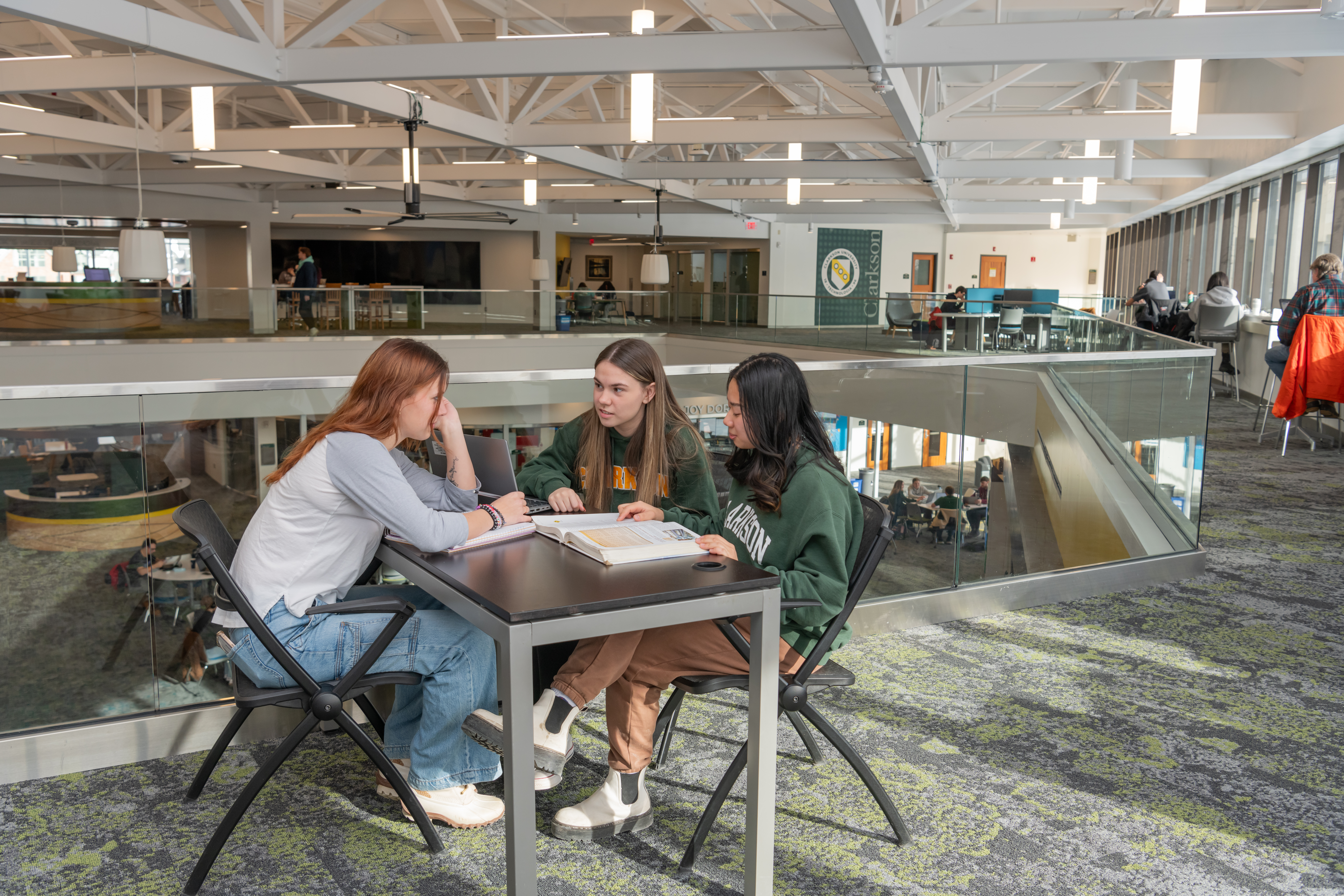 Three female students sit at a table talking with a book and laptop open on the table between them on the second floor balcony of an academic building