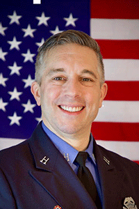 Presenter Rommie Duckworth smiles into the camera with an American flag hanging behind him